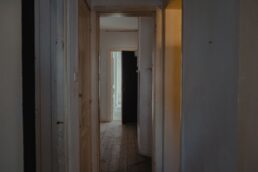 Corridoio lungo e sobrio di una casa vuota con porte chiuse e aperte, luce naturale fioca