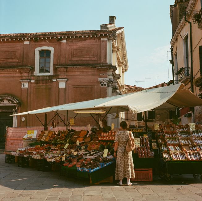Una donna vista di spalle davanti a un banco di frutta e verdura in un mercato italiano all'aperto