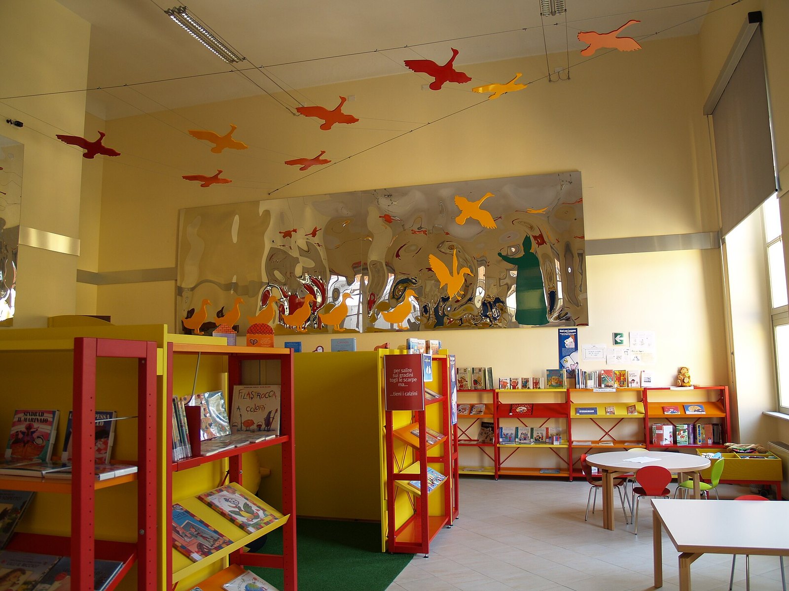 Sala lettura per bambini con scaffali rossi e gialli pieni di libri illustrati e tavolini bassi